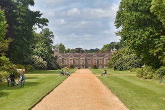 A large, stately mansion with multiple windows sits at the end of a long, sandy path. The surrounding landscape is lush with green grass and various trees and bushes. Several people are walking or sitting on benches along the path and on the grass, enjoying the serene environment. A cloudy sky is visible above the scene.