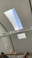 A modern architectural space featuring a ceiling with two elongated skylights allowing natural light to stream in. A sign with bold letters spells out 'TRAVEL IS MY THERAPY' and is prominently displayed across the ceiling. The walls are a smooth off-white with several small, modern wall lights evenly spaced along them. A wooden railing can be seen at the bottom of the image.