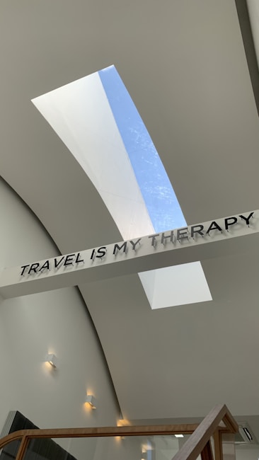 A modern architectural space featuring a ceiling with two elongated skylights allowing natural light to stream in. A sign with bold letters spells out 'TRAVEL IS MY THERAPY' and is prominently displayed across the ceiling. The walls are a smooth off-white with several small, modern wall lights evenly spaced along them. A wooden railing can be seen at the bottom of the image.