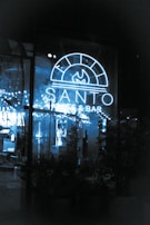 A neon-lit sign reading 'Santo Pizzeria & Bar' illuminates a glass window. The interior appears cozy with string lights reflecting against the glass, greenery visible in the shadows, and a glimpse of the seating area inside.
