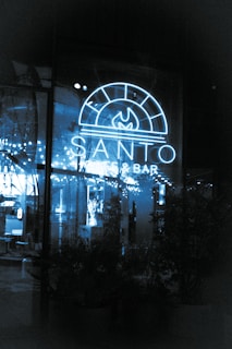 A neon-lit sign reading 'Santo Pizzeria & Bar' illuminates a glass window. The interior appears cozy with string lights reflecting against the glass, greenery visible in the shadows, and a glimpse of the seating area inside.