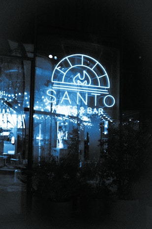 A neon-lit sign reading 'Santo Pizzeria & Bar' illuminates a glass window. The interior appears cozy with string lights reflecting against the glass, greenery visible in the shadows, and a glimpse of the seating area inside.