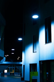 A dimly lit urban alley with modern architecture and wall-mounted circular lamps casting a blue hue over the scene. The buildings appear to be office spaces with large glass windows and minimalistic design. At the bottom right, a sign reads 'C3 bureau' in neon green against a dark background.