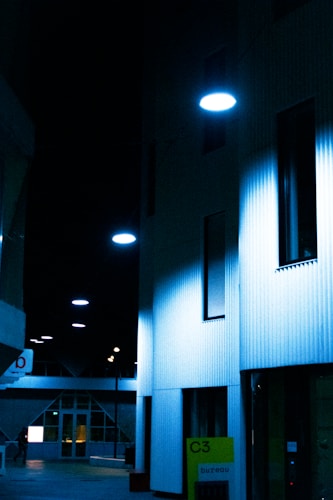 A dimly lit urban alley with modern architecture and wall-mounted circular lamps casting a blue hue over the scene. The buildings appear to be office spaces with large glass windows and minimalistic design. At the bottom right, a sign reads 'C3 bureau' in neon green against a dark background.