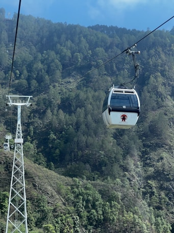 A cable car is suspended from a wire, moving through a mountainous area with dense forest coverage. The tall white support tower is visible alongside the green hills.