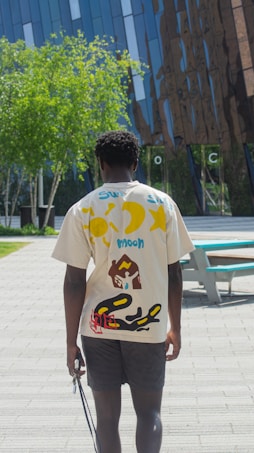 A person with short curly hair is walking on a paved pathway near a modern building. They are wearing a graphic t-shirt with colorful designs including a sun, moon, and other abstract patterns. The background features greenery and reflective glass architecture.