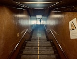 A staircase in a dimly lit underground passageway with tiled walls. The stairs lead up to an illuminated exit where a silhouette of a person is visible. Handrails are present on both sides. There is a caution sign on the right wall providing a warning about slippery conditions when wet.