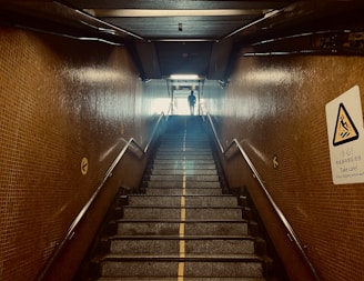 A staircase in a dimly lit underground passageway with tiled walls. The stairs lead up to an illuminated exit where a silhouette of a person is visible. Handrails are present on both sides. There is a caution sign on the right wall providing a warning about slippery conditions when wet.