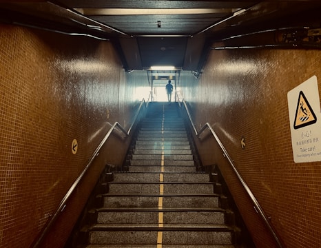 A staircase in a dimly lit underground passageway with tiled walls. The stairs lead up to an illuminated exit where a silhouette of a person is visible. Handrails are present on both sides. There is a caution sign on the right wall providing a warning about slippery conditions when wet.