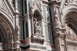 An ornate architectural facade featuring intricate sculptures and detailed stone carvings. The design includes statues of figures set in alcoves, elaborate patterns, and colorful mosaic work. The craftsmanship is highly detailed, reflecting historical and artistic elements.