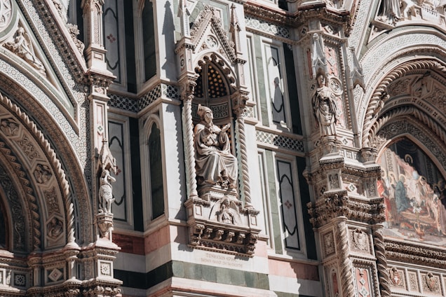 An ornate architectural facade featuring intricate sculptures and detailed stone carvings. The design includes statues of figures set in alcoves, elaborate patterns, and colorful mosaic work. The craftsmanship is highly detailed, reflecting historical and artistic elements.