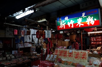 The interior of an Asian market store filled with various goods such as packaged food products, hanging red lanterns, and traditional decorations. A brightly lit sign with Asian characters hangs above, casting colorful light throughout the store. The shelves and floors are packed with boxes and merchandise.