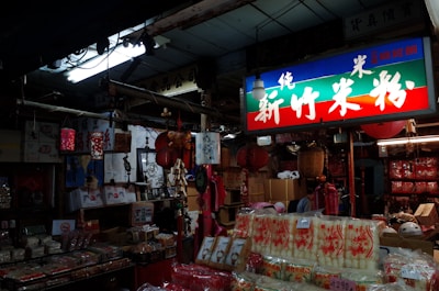 The interior of an Asian market store filled with various goods such as packaged food products, hanging red lanterns, and traditional decorations. A brightly lit sign with Asian characters hangs above, casting colorful light throughout the store. The shelves and floors are packed with boxes and merchandise.