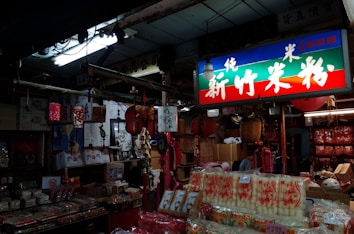 The interior of an Asian market store filled with various goods such as packaged food products, hanging red lanterns, and traditional decorations. A brightly lit sign with Asian characters hangs above, casting colorful light throughout the store. The shelves and floors are packed with boxes and merchandise.