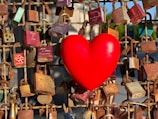 A large red heart is prominently displayed among numerous padlocks attached to a metal fence, symbolizing love and commitment. The padlocks vary in color and condition, some appearing rusty, with names and dates inscribed on them.