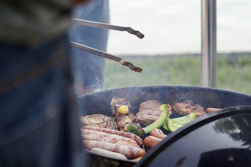 A barbecue in progress with various meats and vegetables sizzling on the grill. Sausages, steaks, and green pepper slices are visible, with a pair of tongs hovering above the food. The grill is slightly open, and there is a blurry foreground of a person beside the grill.