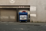 A blue recycling bin is positioned in front of a garage door marked with several signs, including a no entry symbol and warnings for vehicles taller than 1.79 meters and no LPG. The concrete wall and sidewalk show signs of wear and small plants are growing through the cracks.