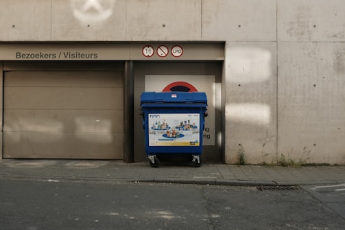 A blue recycling bin is positioned in front of a garage door marked with several signs, including a no entry symbol and warnings for vehicles taller than 1.79 meters and no LPG. The concrete wall and sidewalk show signs of wear and small plants are growing through the cracks.
