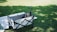 A small dog is comfortably seated in a white and black foldable cart on a grassy field. Sunlight creates dappled shadows on the ground. A white blanket or mat lies beside the cart, partially visible.