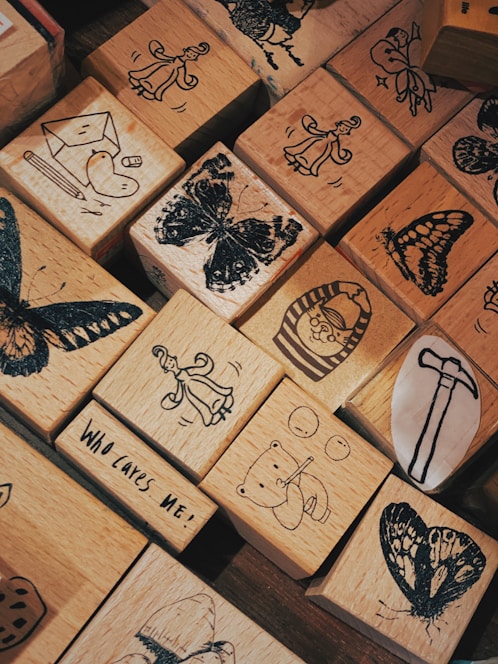 A collection of wooden blocks with various black-ink illustrations. The blocks feature designs such as butterflies, a person in a flowing dress, a bear playing an instrument, and other whimsical drawings. One block includes the text 'Who Cares Me!'.