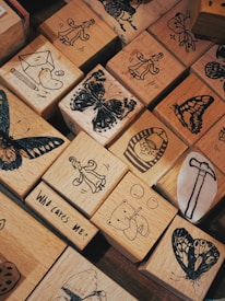 A collection of wooden blocks with various black-ink illustrations. The blocks feature designs such as butterflies, a person in a flowing dress, a bear playing an instrument, and other whimsical drawings. One block includes the text 'Who Cares Me!'.