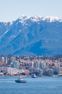 A vibrant cityscape sits beneath a backdrop of towering, snow-capped mountains under a clear blue sky. Tall buildings line the waterfront, while ferries glide across the calm waters.