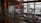 A spacious indoor area with large windows providing natural light, featuring a market or exhibition setup with various stalls and people browsing. The architecture includes wooden elements and modern art installations. The view outside shows some trees and buildings.