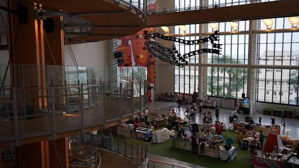 A spacious indoor area with large windows providing natural light, featuring a market or exhibition setup with various stalls and people browsing. The architecture includes wooden elements and modern art installations. The view outside shows some trees and buildings.