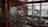 A spacious indoor area with large windows providing natural light, featuring a market or exhibition setup with various stalls and people browsing. The architecture includes wooden elements and modern art installations. The view outside shows some trees and buildings.