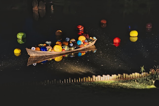 A wooden boat filled with colorful, patterned spheres floats on a dark body of water. More spheres, each uniquely designed, are scattered around the boat. The water reflects these vibrant colors, creating an artistic and surreal effect. In the foreground, a neatly arranged line of small wooden posts is illuminated by artificial light, casting soft shadows on the surrounding grass.