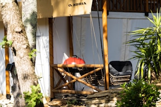 A cozy outdoor setting features a cushioned wicker chair and a red barbecue grill set against a natural backdrop. The scene is partially shaded by trees, and the dappled sunlight adds a tranquil ambiance.