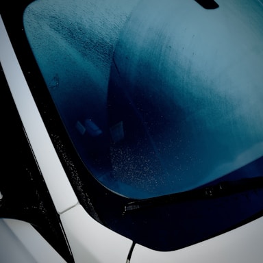 A close-up view of a car windshield shows condensation. The glass has a layer of moisture, partially wiped away, creating an abstract pattern. The view through the window is obscured, with reflections adding to the visual complexity. A black windshield wiper is visible below, in contrast to the smooth white edges of the car.