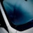 A close-up view of a car windshield shows condensation. The glass has a layer of moisture, partially wiped away, creating an abstract pattern. The view through the window is obscured, with reflections adding to the visual complexity. A black windshield wiper is visible below, in contrast to the smooth white edges of the car.