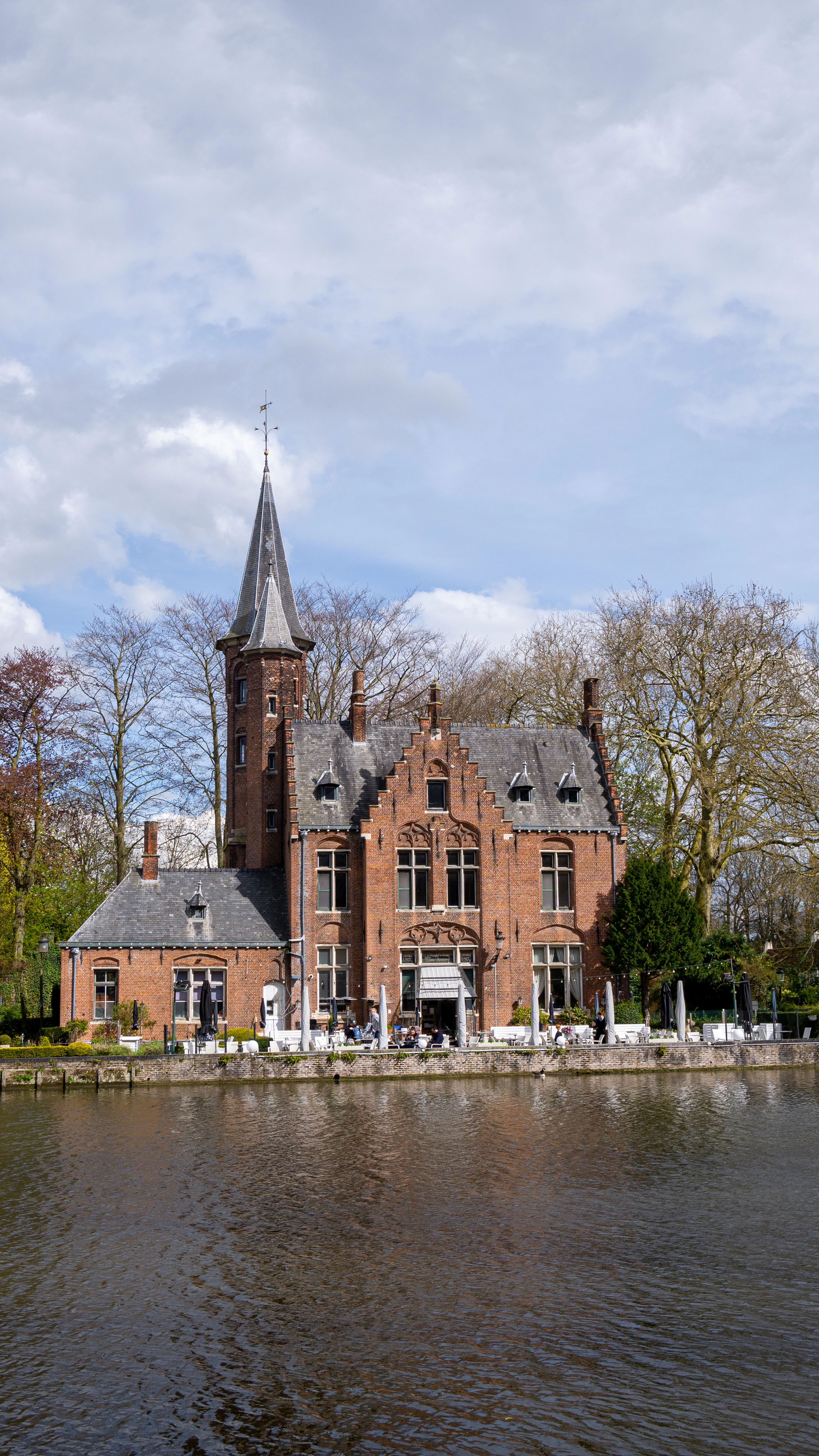 The red-brick turreted castle on the edge of the Minnewater in Bruges, its lakeside terrace set with white parasols and diners beneath bare spring trees and a wide, cloud-streaked sky.