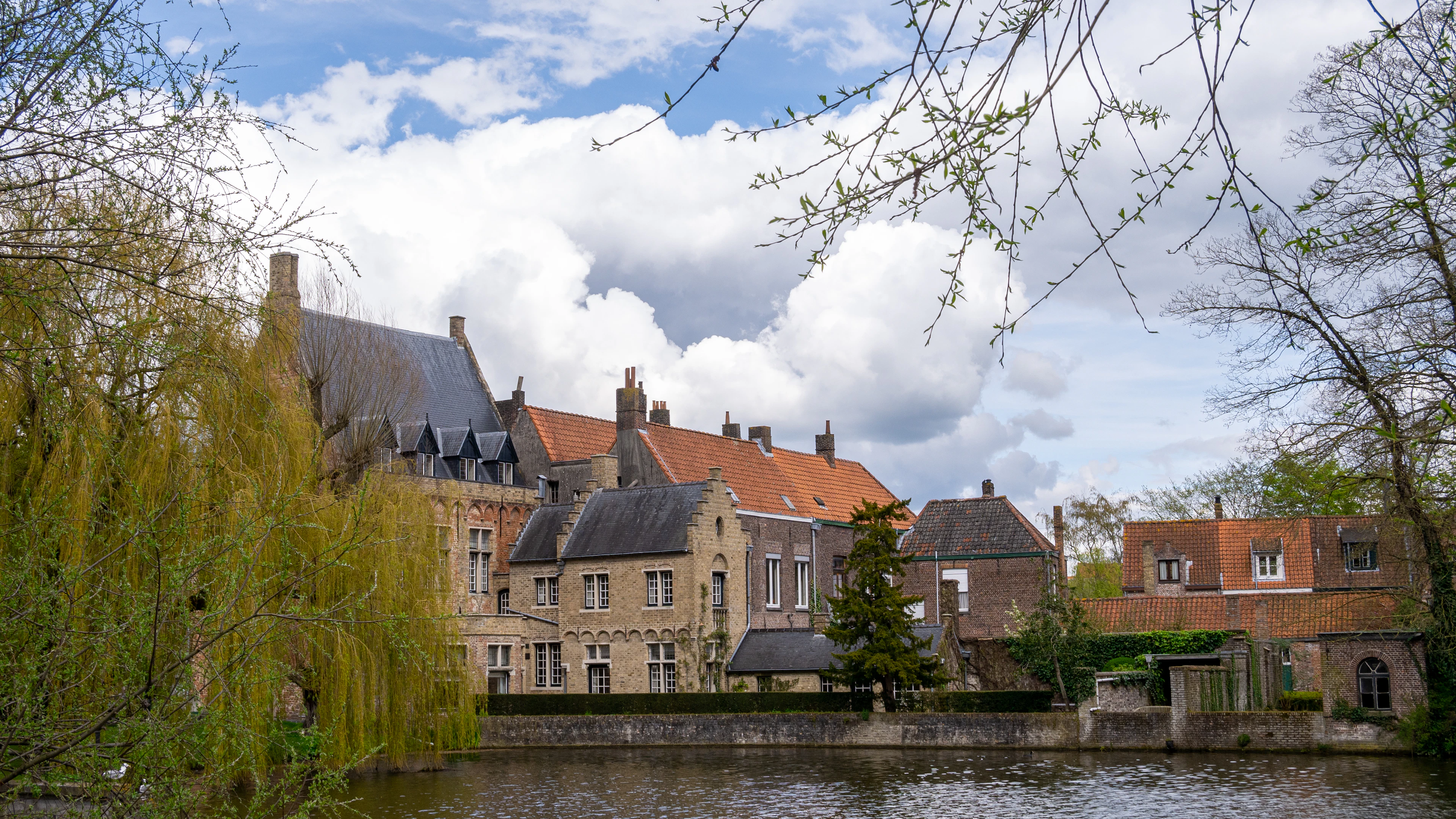 Weeping willow branches frame a canal-side row of brick gabled buildings under a partly cloudy sky.