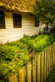 A rustic wooden house with a thatched roof is partially visible. There are two windows with dark frames on the side of the house. In front of the house, a lush, green garden with various plants and small pink flowers thrives behind a wooden picket fence. A tree with purple flowers adds color to the scene.