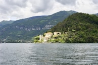 A picturesque villa is nestled amidst lush greenery on a small peninsula surrounded by a serene body of water. The verdant terrain rises into tree-covered hills under a cloudy sky, creating a tranquil and secluded atmosphere.