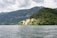 A picturesque villa is nestled amidst lush greenery on a small peninsula surrounded by a serene body of water. The verdant terrain rises into tree-covered hills under a cloudy sky, creating a tranquil and secluded atmosphere.