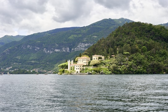A picturesque villa is nestled amidst lush greenery on a small peninsula surrounded by a serene body of water. The verdant terrain rises into tree-covered hills under a cloudy sky, creating a tranquil and secluded atmosphere.