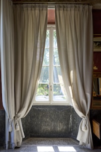 A set of light beige curtains are elegantly draped on either side of a window, allowing soft natural light to pass through. The window offers a view of a garden with green foliage visible outside. The interior features a stone wall with a dark marble texture and a speckled tile floor. Subtle decorative detailing around the window frame adds to the classic look.
