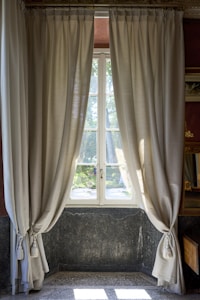 A set of light beige curtains are elegantly draped on either side of a window, allowing soft natural light to pass through. The window offers a view of a garden with green foliage visible outside. The interior features a stone wall with a dark marble texture and a speckled tile floor. Subtle decorative detailing around the window frame adds to the classic look.