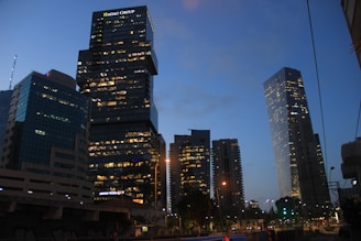 A cityscape at dusk features several modern skyscrapers with their windows illuminated, creating a contrast against the deepening blue sky. The skyline includes a prominent building displaying 'HAGAG GROUP' at the top. Streetlights and smaller buildings are visible in the foreground, adding to the urban atmosphere.