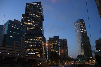 A cityscape at dusk features several modern skyscrapers with their windows illuminated, creating a contrast against the deepening blue sky. The skyline includes a prominent building displaying 'HAGAG GROUP' at the top. Streetlights and smaller buildings are visible in the foreground, adding to the urban atmosphere.