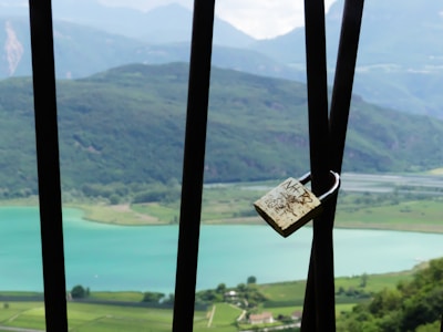 A padlock is attached to black metal bars overlooking a serene landscape featuring a turquoise lake, greenery, and distant mountains under a partly cloudy sky.