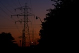 Silhouettes of power lines and electrical towers are set against a twilight sky, with a gradient of deep blue transitioning to a warm orange near the horizon. The surrounding trees and structures are in shadow, creating a stark contrast with the vibrant sky.