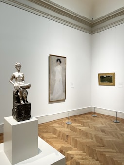 A gallery room featuring a silver sculpture on a pedestal, a portrait of a woman in a long white dress, and a landscape painting with earthy tones. The walls are white, and the floor is made of herringbone-patterned wood.