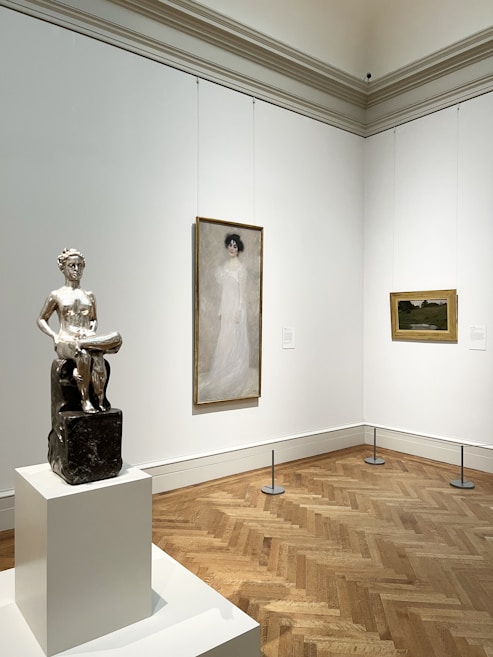 A gallery room featuring a silver sculpture on a pedestal, a portrait of a woman in a long white dress, and a landscape painting with earthy tones. The walls are white, and the floor is made of herringbone-patterned wood.