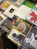 A selection of magazines arranged on a table, with various covers visible. The magazines feature bold, colorful designs and photography; some include images of smiling people and artistic illustrations. Texts on the covers are diverse, with some emphasizing topics like textiles, interviews, and reading material. The overall presentation has a casual yet organized aesthetic.