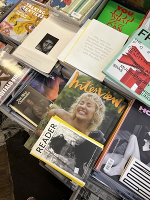 A selection of magazines arranged on a table, with various covers visible. The magazines feature bold, colorful designs and photography; some include images of smiling people and artistic illustrations. Texts on the covers are diverse, with some emphasizing topics like textiles, interviews, and reading material. The overall presentation has a casual yet organized aesthetic.