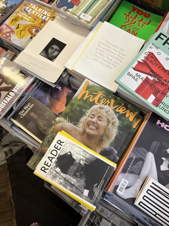 A selection of magazines arranged on a table, with various covers visible. The magazines feature bold, colorful designs and photography; some include images of smiling people and artistic illustrations. Texts on the covers are diverse, with some emphasizing topics like textiles, interviews, and reading material. The overall presentation has a casual yet organized aesthetic.